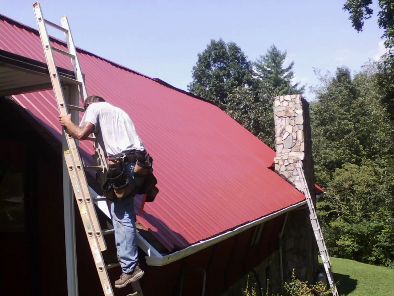 Roofer on a ladder beside a red metal roof and stone chimney — Soffit & Fascia Repair by Roofing Brothers in Mahanoy City